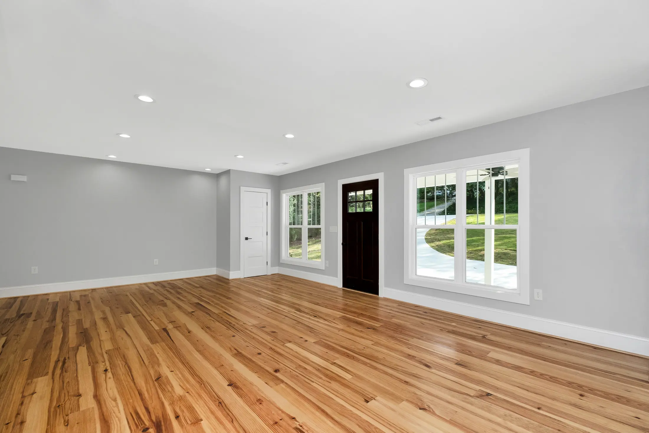 Empty apartment living room with warm oak plank flooring, grey walls, a window and a dark front door